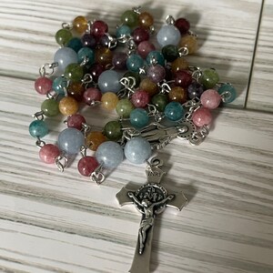 May include: A multi-colored stone rosary with a silver crucifix featuring a figure of Jesus Christ. The crucifix has the inscription "INRI" on the top.