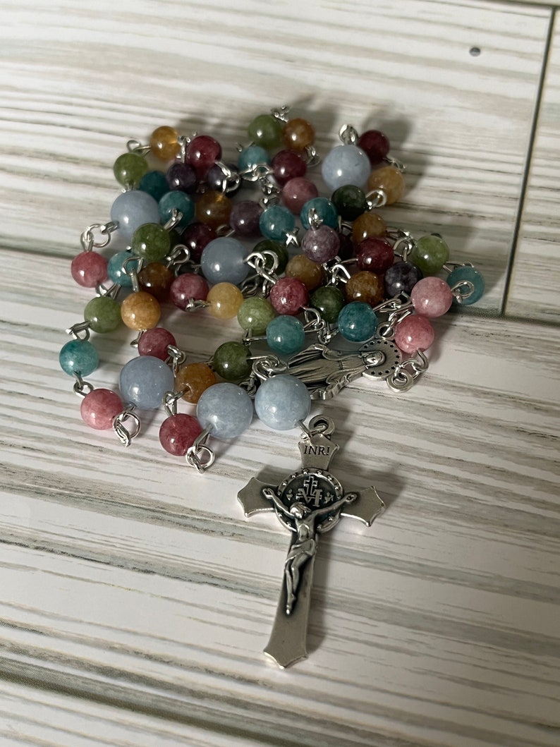 May include: A multi-colored stone rosary with a silver crucifix featuring a figure of Jesus Christ. The crucifix has the inscription "INRI" on the top.