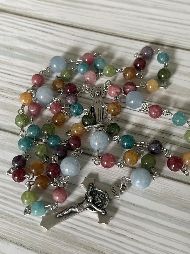 May include: A colorful rosary with a silver crucifix and a silver charm of the Virgin Mary. The beads are made of various shades of blue, green, brown, and pink.