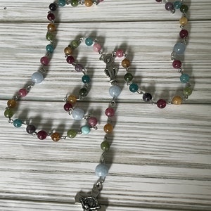 May include: A multi-colored gemstone rosary with a silver crucifix and a silver charm of the Virgin Mary. The rosary beads are made of various shades of blue, green, brown, and pink.