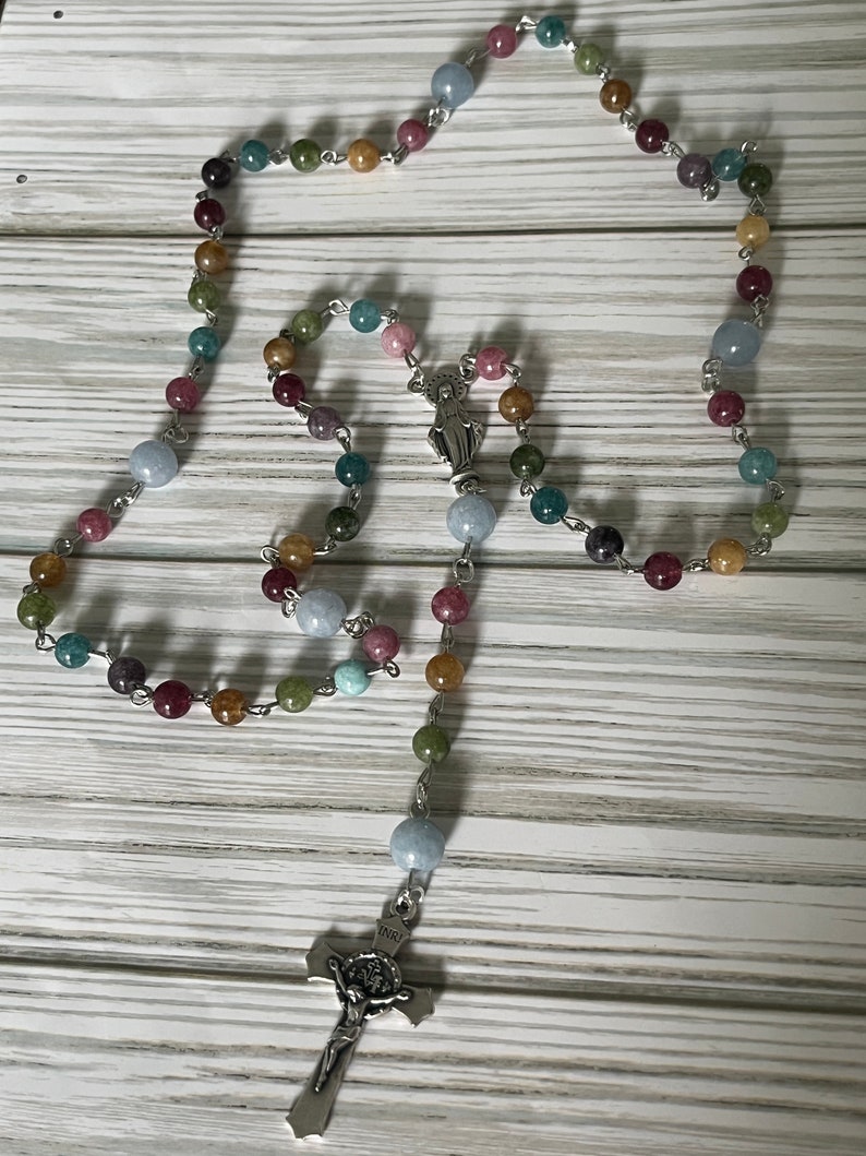 May include: A multi-colored gemstone rosary with a silver crucifix and a silver charm of the Virgin Mary. The rosary beads are made of various shades of blue, green, brown, and pink.