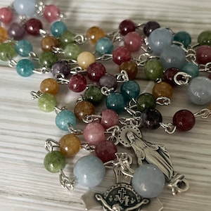 May include: A silver rosary with a silver crucifix and a silver charm of the Virgin Mary. The beads are made of various colored stones, including blue, green, pink, and brown.