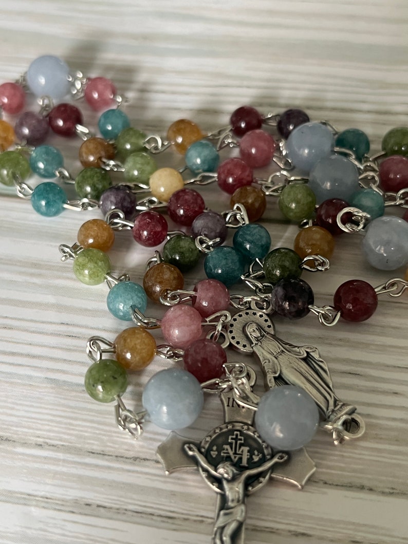 May include: A silver rosary with a silver crucifix and a silver charm of the Virgin Mary. The beads are made of various colored stones, including blue, green, pink, and brown.