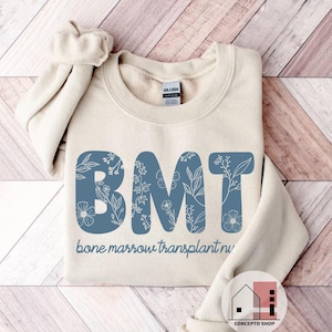 May include: A beige sweatshirt with the letters "BMT" in a blue floral design. The words "bone marrow transplant nurse" are printed below the letters. The sweatshirt is folded on a light wood-grain surface.