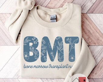 May include: A beige sweatshirt with the letters "BMT" in a blue floral design. The words "bone marrow transplant nurse" are printed below the letters. The sweatshirt is folded on a light wood-grain surface.