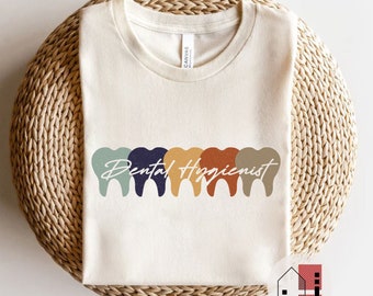 May include: A beige t-shirt with the text "Dental Hygienist" in a colourful font. The text is surrounded by five colourful tooth outlines.