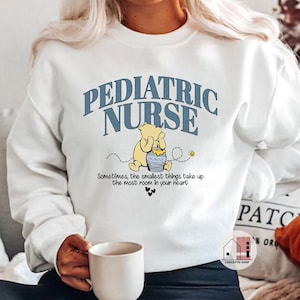 May include: White sweatshirt with blue text that reads "Pediatric Nurse" and a cartoon Winnie the Pooh holding a honey pot. The text below the image reads "Sometimes, the smallest things take up the most room in your heart."