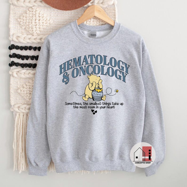 May include: A light gray sweatshirt with the words "HEMATOLOGY & ONCOLOGY" in blue arched text. Below is a cartoon of Winnie the Pooh with a honey pot. The text "Sometimes, the smallest things take up the most room in your heart" is also printed.