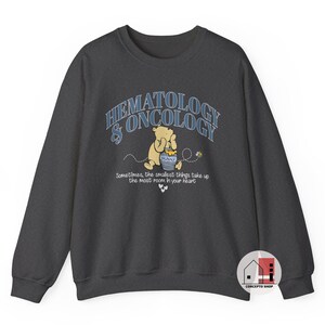 May include: Dark gray sweatshirt with the words "HEMATOLOGY & ONCOLOGY" in light blue arched text. A cartoon bear is holding a honey pot. The text "Sometimes, the smallest things take up the most room in your heart" is below.
