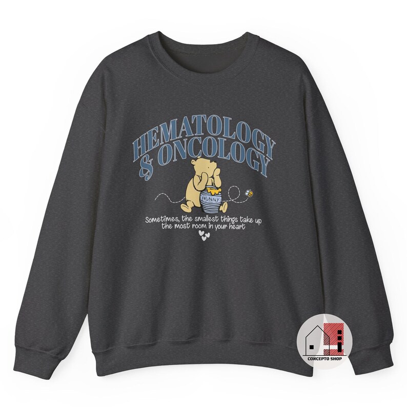 May include: Dark gray sweatshirt with the words "HEMATOLOGY & ONCOLOGY" in light blue arched text. A cartoon bear is holding a honey pot. The text "Sometimes, the smallest things take up the most room in your heart" is below.