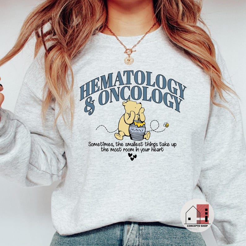 May include: A light gray sweatshirt featuring the text "HEMATOLOGY & ONCOLOGY" in blue. Below the text is a cartoon bear holding a honey pot. The sweatshirt also has the quote, "Sometimes, the smallest things take up the most room in your heart."