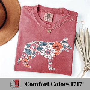 Pu&ograve; includere: Una maglietta rossa con una stampa floreale a forma di silhouette di pastore tedesco. La maglietta ha un'etichetta che dice "Comfort Colors 1717".