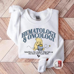 May include: White sweatshirt with the words "HEMATOLOGY & ONCOLOGY" in blue lettering. A cartoon bear is holding a honey pot. The text "Sometimes, the smallest things take up the most room in your heart" is below the bear.
