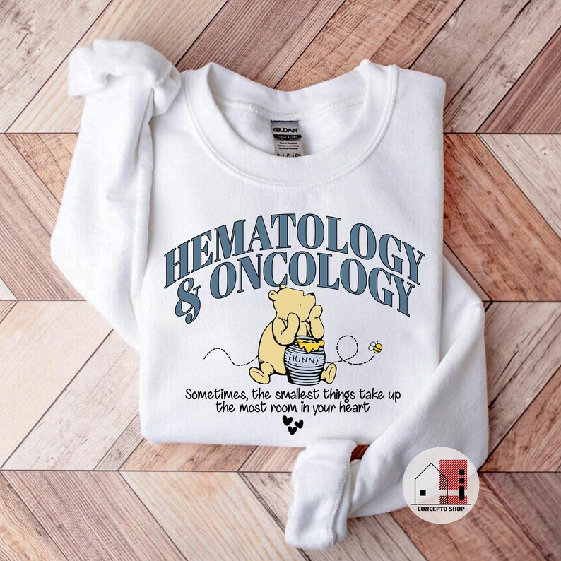 May include: White sweatshirt with the words "HEMATOLOGY & ONCOLOGY" in blue lettering. A cartoon bear is holding a honey pot. The text "Sometimes, the smallest things take up the most room in your heart" is below the bear.