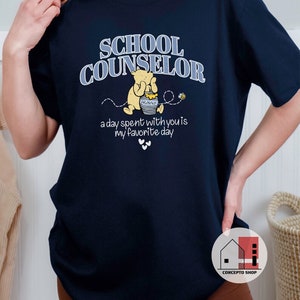 May include: Navy blue t-shirt with the text "School Counselor" in white and blue lettering. A cartoon Winnie the Pooh bear is holding a jar of honey with the text "Hunny" on it. The text "a day spent with you is my favorite day" is below the bear.