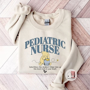 May include: A beige sweatshirt with the text "PEDIATRIC NURSE" in blue lettering. A cartoon Winnie the Pooh character is holding a jar of honey with the text "HUNNY" on it. The text "Sometimes, the smallest things take up the most room in your heart" is below the image.