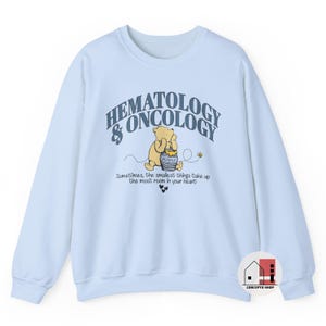 May include: A light blue sweatshirt featuring the text "HEMATOLOGY & ONCOLOGY" above a cartoon illustration of Winnie the Pooh with a honey pot. The text "Sometimes, the smallest things take up the most room in your heart" is below the illustration.