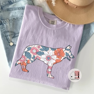 Pu&ograve; includere: Una maglietta color lavanda con una stampa floreale a forma di silhouette di pastore tedesco. La maglietta ha il testo "Comfort Colors" sull'etichetta del collo e "Concepto Shop" su una piccola icona di casa in basso a destra.