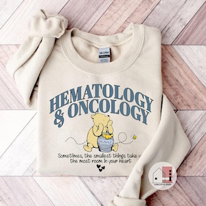 May include: Cream-colored sweatshirt with the words "HEMATOLOGY & ONCOLOGY" in blue lettering. A cartoon bear is seated next to a honey pot. The text "Sometimes, the smallest things take the most room in your heart" is below the bear.
