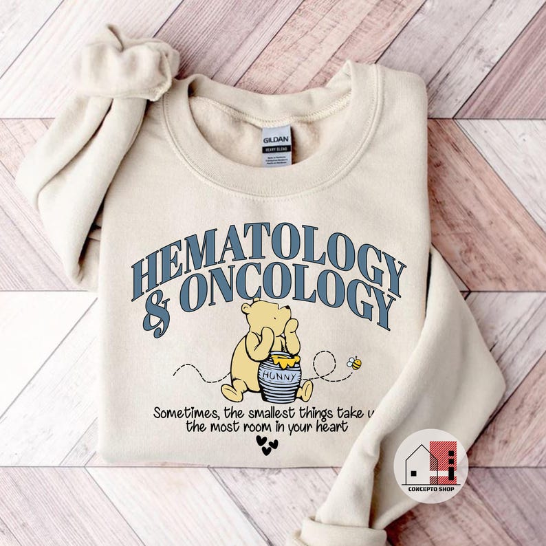 May include: Cream-colored sweatshirt with the words "HEMATOLOGY & ONCOLOGY" in blue lettering. A cartoon bear is seated next to a honey pot. The text "Sometimes, the smallest things take the most room in your heart" is below the bear.