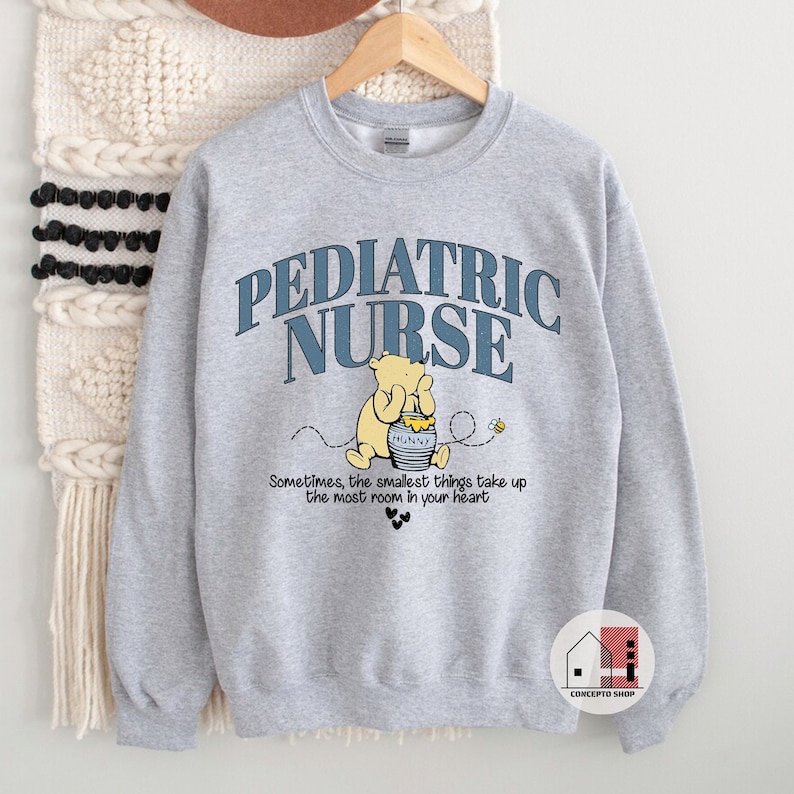 May include: Gray sweatshirt with the text "PEDIATRIC NURSE" in blue and white lettering. A cartoon Winnie the Pooh character is holding a jar of honey with the word "HUNNY" on it. The text "Sometimes, the smallest things take up the most room in your heart" is below the image.