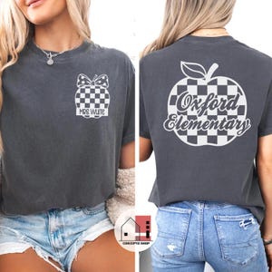 May include: Dark gray t-shirt with a checkered apple design on the back and a checkered apple with a bow on the front. The front of the shirt has the text "MRS. WHITE". The back of the shirt has the text "Oxford Elementary".