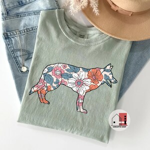 Pu&ograve; includere: Una maglietta verde chiaro con una silhouette di cane con motivo floreale. Il cane &egrave; rivolto a destra e presenta un motivo floreale rosa, bianco e blu. La maglietta ha un'etichetta che dice "Comfort Colors".