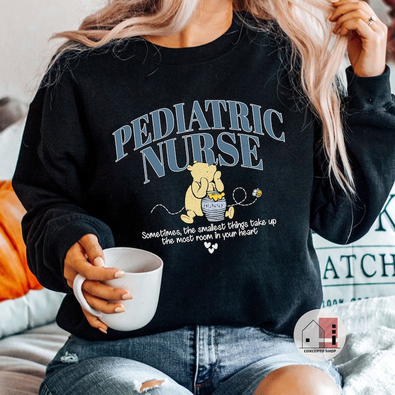 May include: Black sweatshirt with the text "PEDIATRIC NURSE" in blue lettering. A cartoon Winnie the Pooh character is holding a jar of honey with the text "HUNNY". The text "Sometimes, the smallest things take up the most room in your heart" is below the cartoon.