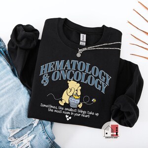 May include: Black sweatshirt with the words "HEMATOLOGY & ONCOLOGY" in blue and white. A cartoon bear is holding a honey pot. The text "Sometimes, the smallest things take up the most room in your heart" is below the bear. A silver necklace is on top of the sweatshirt.