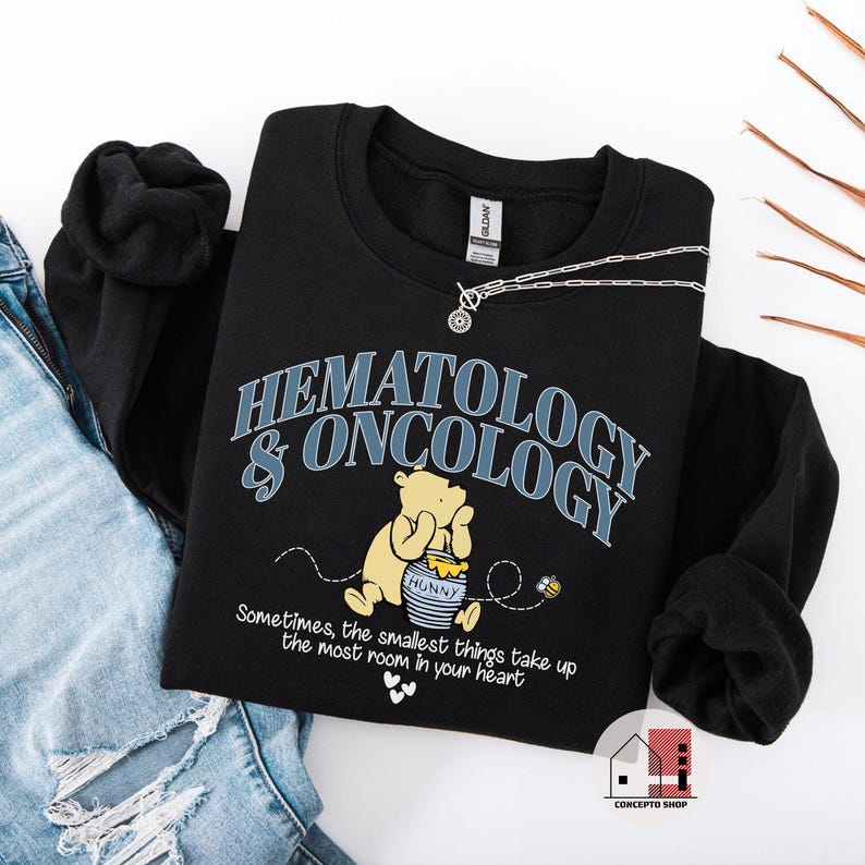 May include: Black sweatshirt with the words "HEMATOLOGY & ONCOLOGY" in blue and white. A cartoon bear is holding a honey pot. The text "Sometimes, the smallest things take up the most room in your heart" is below the bear. A silver necklace is on top of the sweatshirt.