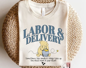 May include: Cream-colored t-shirt with the text "LABOR & DELIVERY" in blue. A cartoon bear is holding a honey pot with the word "HUNNY" on it. The shirt also has the quote "Sometimes, the smallest things take up the most room in your heart."