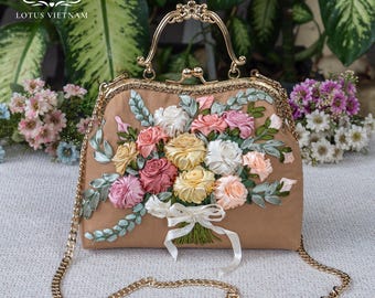 Könnte beinhalten: Eine hellbraune Handtasche mit einem goldfarbenen Metallrahmen, einem Tragegriff und einem abnehmbaren Kettenriemen. Die Vorderseite ist mit einem dreidimensionalen Bandstickerei-Bouquet aus Rosen in Rosa, Gelb und Weiß sowie grünen Blättern verziert.