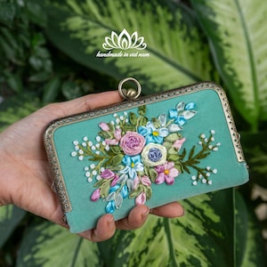 May include: A teal clutch bag with a floral embroidery design. The bag features pink, blue, and white flowers with green leaves. The frame is bronze-coloured, and the text "handmade in viet nam" is visible.