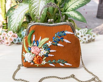 May include: A brown velvet clutch bag with a floral embroidery design. The bag has a metal clasp closure and a chain strap.