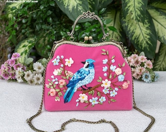 Könnte beinhalten: Eine pinkfarbene Handtasche mit Metallverschluss und Griff, bestickt mit einem blauen Vogel auf einem Ast mit Blumen. Die Tasche hat einen Kettenriemen und den Text "handmade in viet nam".