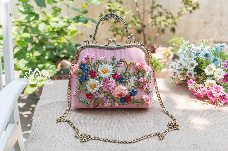 May include: A pink satin clutch bag with a floral embroidery design. The bag has a gold chain strap and a metal clasp closure.