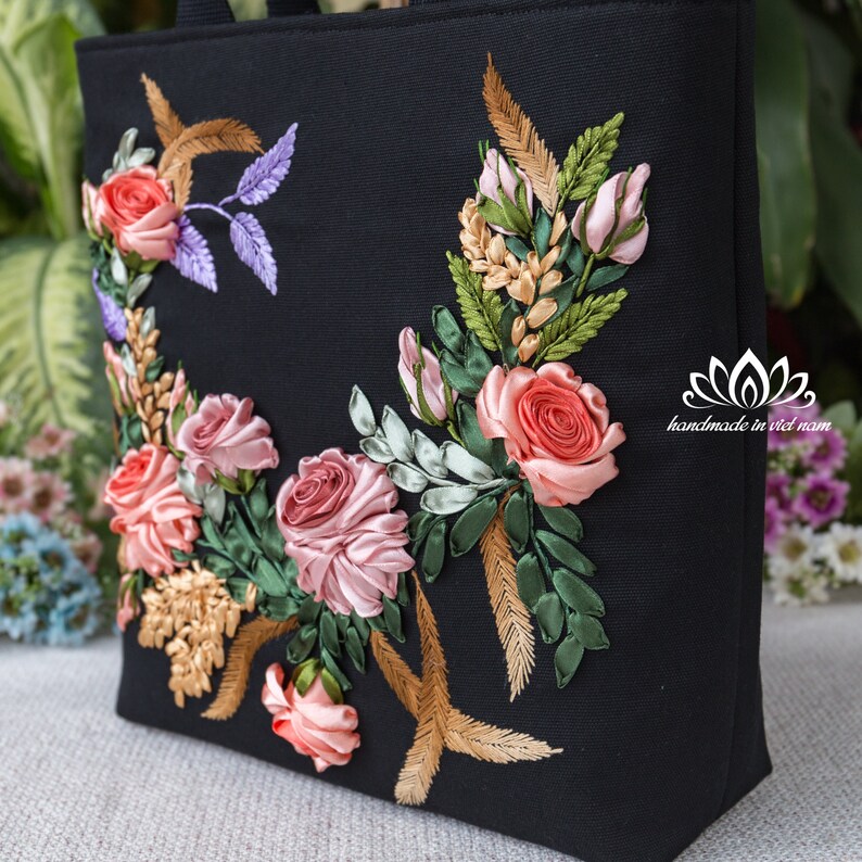 K&ouml;nnte beinhalten: Schwarze Canvas-Tragetasche mit einem floralen Design, das mit rosa, rotem und goldenem Band bestickt ist. Die Stickerei zeigt Rosen, Bl&auml;tter und Federn. Die Tasche tr&auml;gt den Text "handmade in viet nam" in der unteren rechten Ecke.