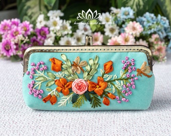 May include: A teal blue fabric clutch purse with a floral embroidery design. The embroidery features orange, pink, and green ribbon flowers, leaves, and a dragonfly.