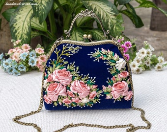 Può includere: Una pochette in velluto blu con una tracolla a catena dorata e ricamo di rose in nastro rosa. La borsa ha una chiusura a scatto e un manico decorativo.