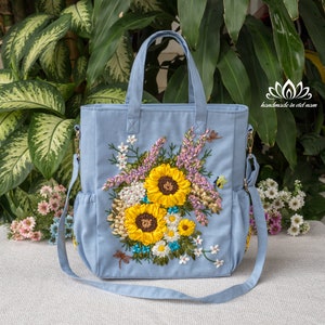 May include: A light blue tote bag with a floral design embroidered with ribbon. The design features sunflowers, daisies, and other flowers in shades of yellow, white, and pink. The bag has a long strap and a top handle.