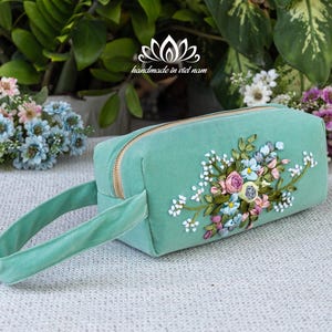 May include: A rectangular, mint green cosmetic bag with a gold zipper and a matching handle. The bag is adorned with a floral embroidery design in pastel colors. The text "handmade in viet nam" is visible.