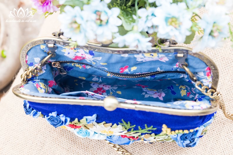 May include: A blue velvet clutch purse with a floral design and a gold chain strap. The purse is open and the inside is lined with a blue and white floral fabric.