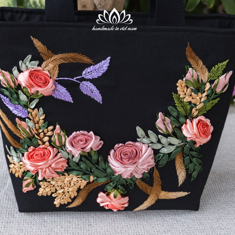 K&ouml;nnte beinhalten: Schwarze Tragetasche mit einem floralen Design, das mit rosa, pfirsichfarbenem und gr&uuml;nem Band bestickt ist. Der Text "handmade in viet nam" ist auf die Tasche gedruckt.