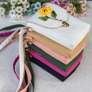 May include: A stack of six colourful fabric phone pouches with a white pouch on top featuring an embroidered sunflower design. The pouches have a flap closure with a gold metal clasp. The pouches are handmade in Vietnam.