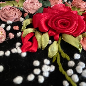 Op de afbeelding: Close-up van een zwarte stof met rode, roze en groene lintborduursels. De borduursels tonen rozen en bladeren. De stof is bedekt met kleine witte stippen.