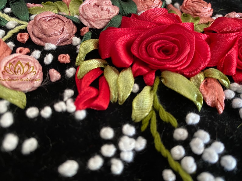 Op de afbeelding: Close-up van een zwarte stof met rode, roze en groene lintborduursels. De borduursels tonen rozen en bladeren. De stof is bedekt met kleine witte stippen.