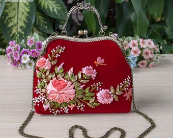 Könnte beinhalten: Eine tiefrote Samthandtasche mit einem verzierten Rahmen aus antikem Messing und einem passenden Ketten-Schulterriemen. Die Vorderseite ist mit dreidimensionaler Bandstickerei aus rosa Rosen, grünen Blättern und einer kleinen orangefarbenen Libelle verziert.