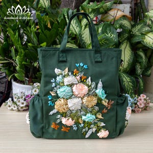 以下が含まれることがあります： 濃い緑色のキャンバス地のトートバッグに、大きな刺繍の花柄が施されています。花はリボンで作られており、ピンク、ブルー、ホワイト、グリーンの色合いです。