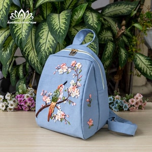 Peut inclure: Un sac à dos en toile bleu clair avec une broderie d'oiseau et de fleurs. La broderie est réalisée dans des tons de rose, blanc, vert et orange.