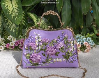 May include: A lavender satin handbag with a gold chain strap and a gold clasp. The bag is decorated with embroidered flowers, including roses and lilies.